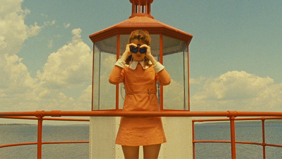 Scene from Wes Anderson film with woman holding binoculars on top of lighthouse wearing orange